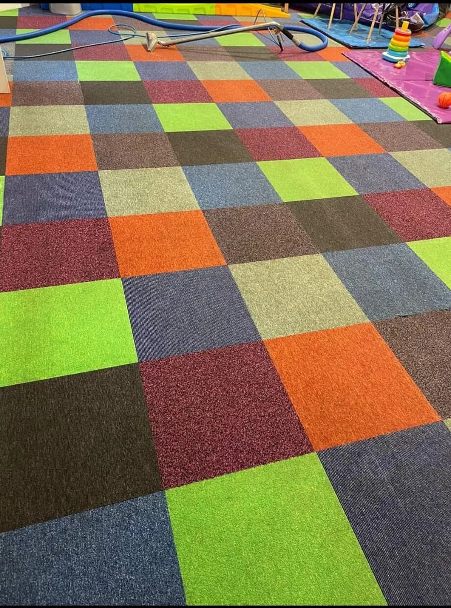 A floor of multicoloured commercial carpet tiles after cleaning, with the green, orange and blue tiles showing bright, even colour and an extraction hose still on the floor