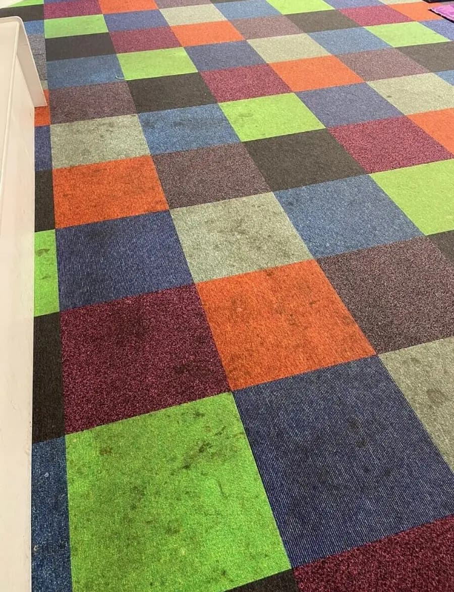 A floor of multicoloured commercial carpet tiles with grey soiling worn into the green and pale tiles, photographed before cleaning