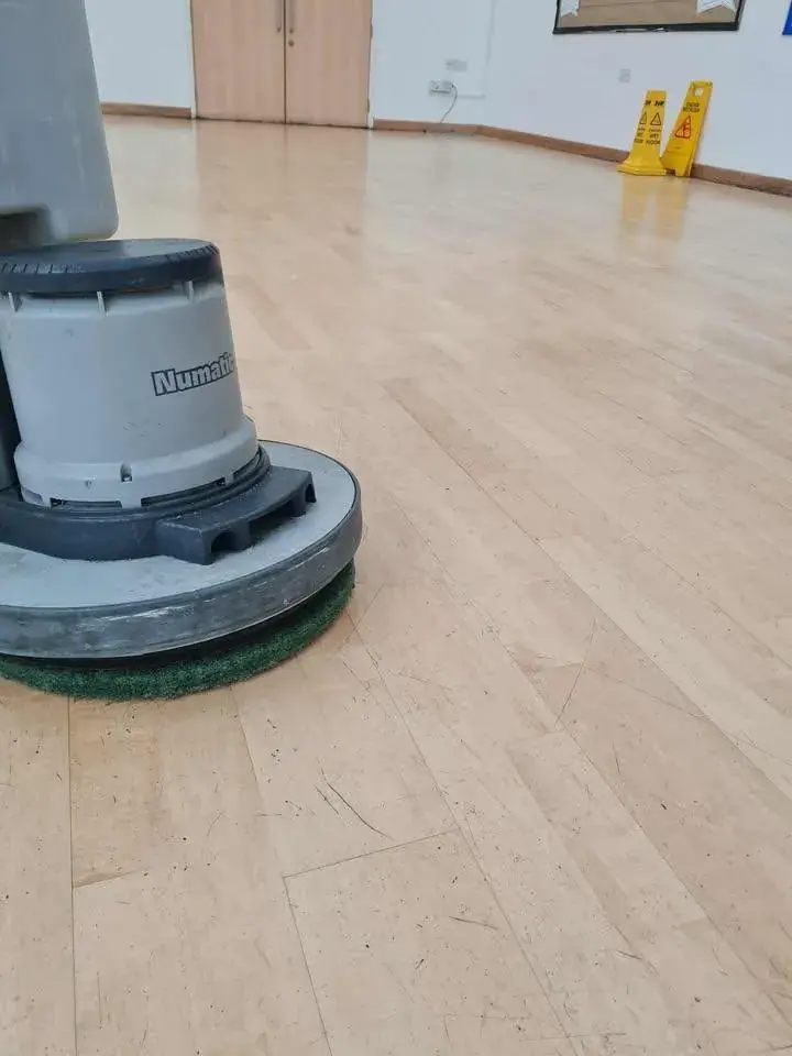 A rotary floor cleaning machine part way along a pale wood effect hard floor in an empty room